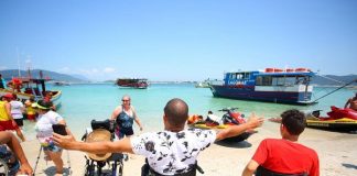 homem em cadeira de rodas com os braços abertos na beira da praia da ilha do campeche e outras pessoas, em pé ou cadeiras de rodas, em volta; barcos na água