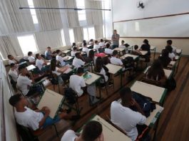 sala de aula cheia, com estudantes sentados nas carteiras e professor à frente em pé