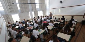sala de aula cheia, com estudantes sentados nas carteiras e professor à frente em pé