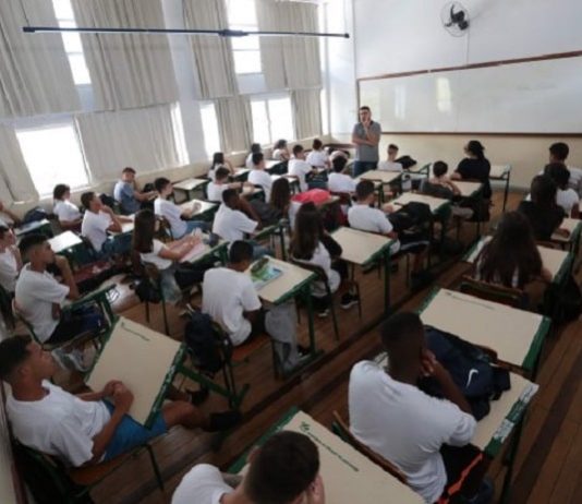 sala de aula cheia, com estudantes sentados nas carteiras e professor à frente em pé