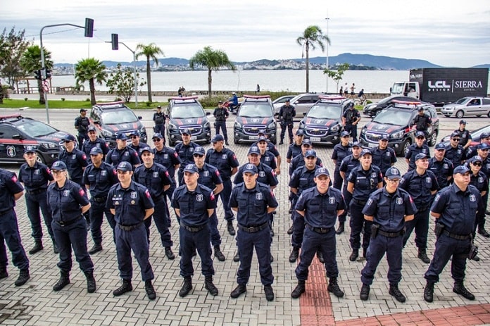 agentes da guarda alinhados em sentido com viaturas ao fundo estacionadas ordenadas em volta, na frente da prefeitura