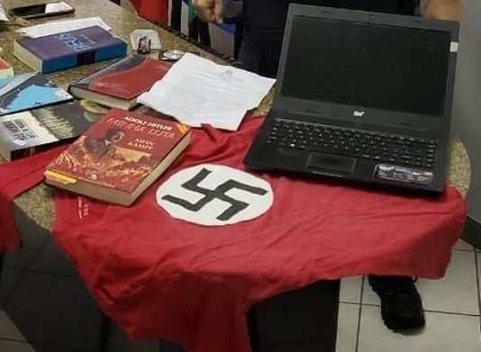 sobre uma mesa de mármore estão diversos livros do nazismo, camiseta com suástica e notebook