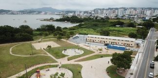 foto aérea do parque, com pista de caminhada, grande gramado mar ao fundo e pequenos apartamentos ao lado