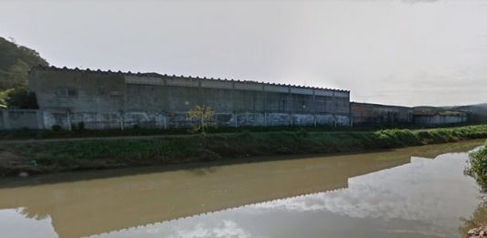 imagem do rio com mato ao redor, pequena rua de terra e prédio na margem oposta