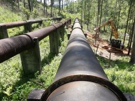 duas grandes tubulações de água em área de mata e inclinada com uma retroescavadeira ao lado
