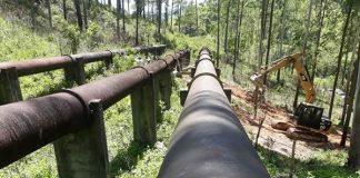 duas grandes tubulações de água em área de mata e inclinada com uma retroescavadeira ao lado