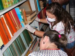 duas mulheres, uma delas usando máscara, mexem em livros de capa dura em prateleiras de estante