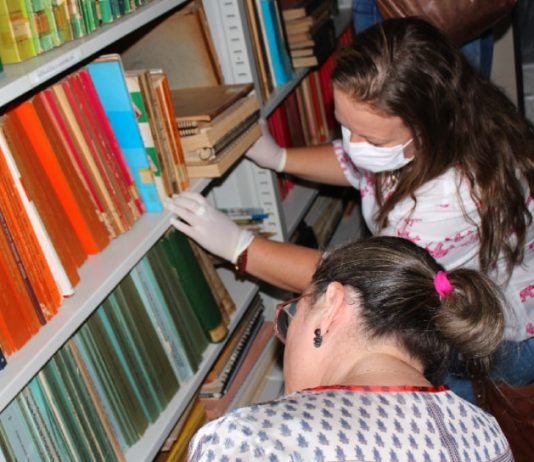 2 milhões de páginas do arquivo público de Santa Catarina serão digitalizadas duas mulheres, uma delas usando máscara, mexem em livros de capa dura em prateleiras de estante