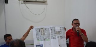 dois homens seguram uma planta baixa de um prédio; um deles está em pé falando ao microfone