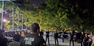 policiais civis reunidos em pé em um estacionamento à noite com árvores em volta ouvindo um deles falar, de costas para a câmera