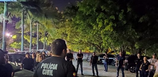 policiais civis reunidos em pé em um estacionamento à noite com árvores em volta ouvindo um deles falar, de costas para a câmera