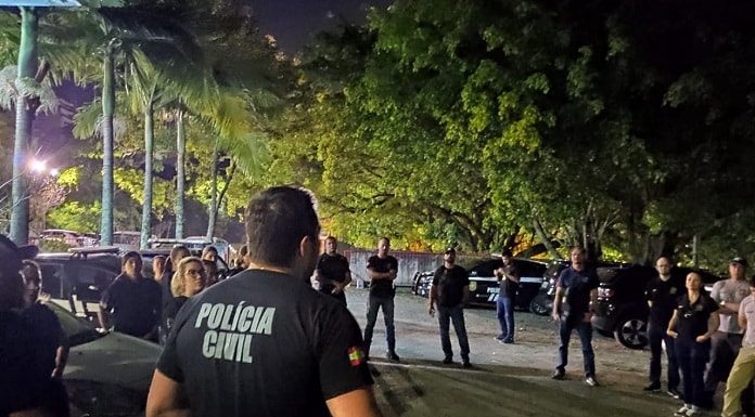 policiais civis reunidos em pé em um estacionamento à noite com árvores em volta ouvindo um deles falar, de costas para a câmera
