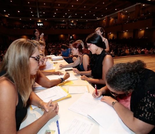 Rede estadual de ensino recebe reforço de 1.842 novos servidores mesa onde mulheres estão dos dois lados, uma assinando papeis, outras entregando