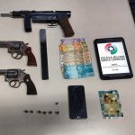 armas, celular e balas dispostas sobre uma mesa com tablet com logo da pmsc