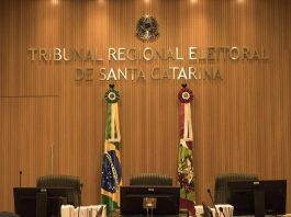 parede decorada em madeira com grande letreiro dourado "Tribunal Regional Eleitoral de Santa Catarina", bandeiras do brasil e sc e grande cadeiras em roda