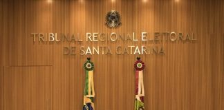parede decorada em madeira com grande letreiro dourado "Tribunal Regional Eleitoral de Santa Catarina", bandeiras do brasil e sc e grande cadeiras em roda