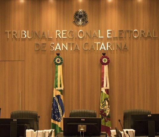 Dois institutos de pesquisa são multados por juiz do TRE parede decorada em madeira com grande letreiro dourado "Tribunal Regional Eleitoral de Santa Catarina", bandeiras do brasil e sc e grande cadeiras em roda