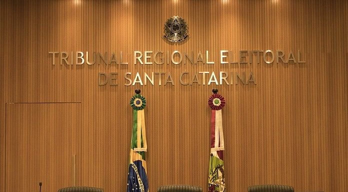 parede decorada em madeira com grande letreiro dourado "Tribunal Regional Eleitoral de Santa Catarina", bandeiras do brasil e sc e grande cadeiras em roda