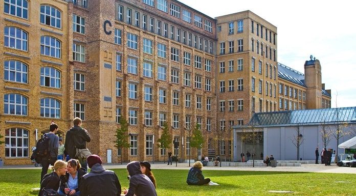 jovens sentados em roda em gramado de uma universidade do exterior, com grande edifício histórico ao fundo