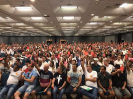 grande saguão com milhares de pessoas sentadas com os braços levantados mostrando um cartão vermelho