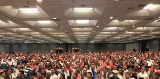 grande saguão com milhares de pessoas sentadas com os braços levantados mostrando um cartão vermelho