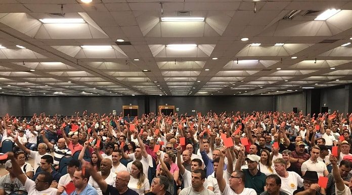 grande saguão com milhares de pessoas sentadas com os braços levantados mostrando um cartão vermelho