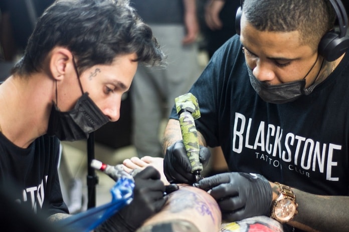 dois tatuadores usando máscara fazendo tatuagem em alguém
