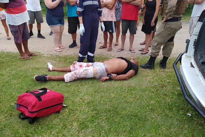 homem caído no chão algemado e ensanguentado em gramado no dia do assassinato com diversas pessoas em volta e policial e paramédica do samu