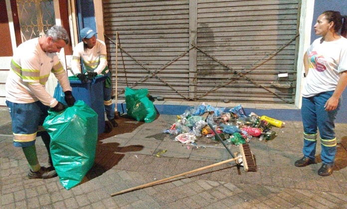 um funcionário e duas funcionárias da comcap recolhem lixo de um calçadão em frente a comércio; homem fecha sacola de lixo