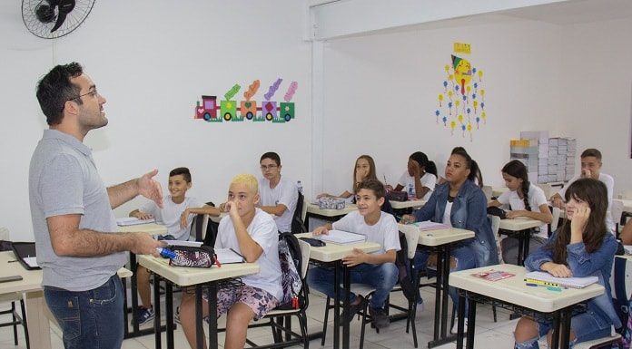 professor fala e gesticula em pé na frente da sala com alunos e alunas sentados nas carteiras