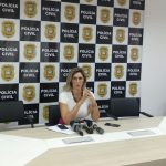 delegada sentada atrás de uma mesa falando e gesticulando e ao fundo painel com logos da polícia civil de sc