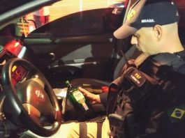 policial da prf sentado dentro de um carro da nissan segurando e olhando para uma garrafa de bebida alcoólica