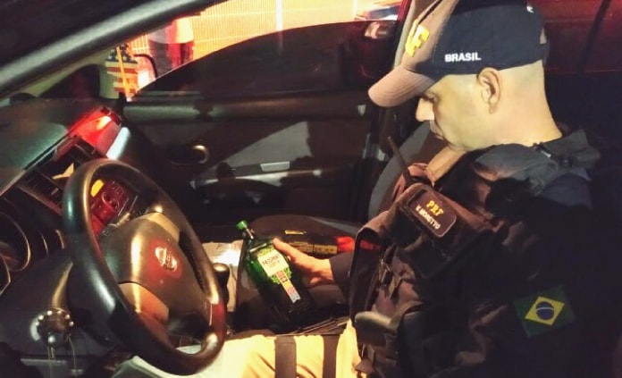 policial da prf sentado dentro de um carro da nissan segurando e olhando para uma garrafa de bebida alcoólica