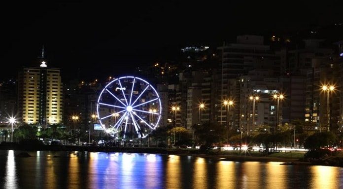 foto noturna da orla da beira mar com roda gigante em destaque e luzes do equipamento e da rua refletidas na água