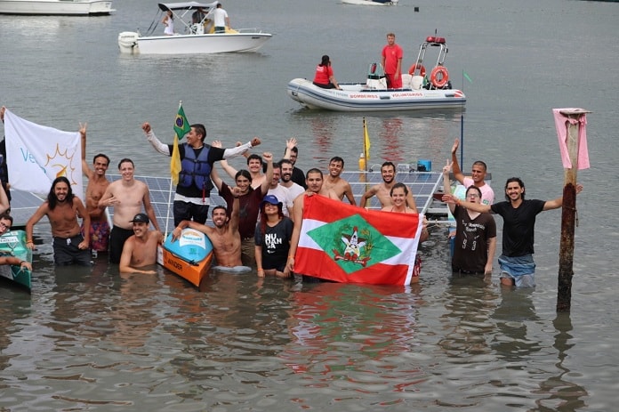jovens na água com bandeira de sc e barcos ao fundo posam comemorando para a foto