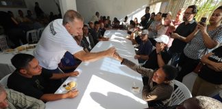 prefeito cumprimenta um homem do outro lado de uma grande mesa de refeitório cheia de gente sentada e em pé em volta