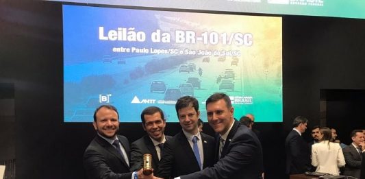 CCR vence leilão da BR 101 Sul com proposta de R$ 1,97 para cada pedágio quatro homens seguram martelo sobre balcão sorrindo para a câmera com telão ao fundo "leilão da br 101 sul sc"