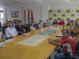grupo de homens e mulheres reunidos em volta de grande mesa em sala que há painel da prefeitura ao fundo