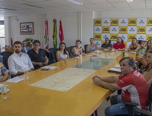 grupo de homens e mulheres reunidos em volta de grande mesa em sala que há painel da prefeitura ao fundo