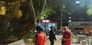 duas assistentes sociais com coletes da prefeitura ao lado de homem deitado com cobertor em banco de praça e guarda ao fundo