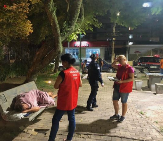 Câmara de São José aprova internação involuntária de dependentes químicos duas assistentes sociais com coletes da prefeitura ao lado de homem deitado com cobertor em banco de praça e guarda ao fundo