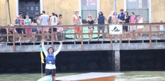 O piloto Marcos comemora o término da prova, de pé no barco em frente ao um pier com pessoas