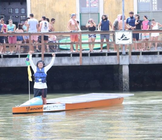 O piloto Marcos comemora o término da prova, de pé no barco em frente ao um pier com pessoas