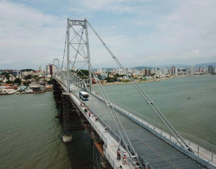 ônibus do transporte coletivo municipal de florianópolis passando pela ponte hercílio luz em foto aérea com o mar abaixo