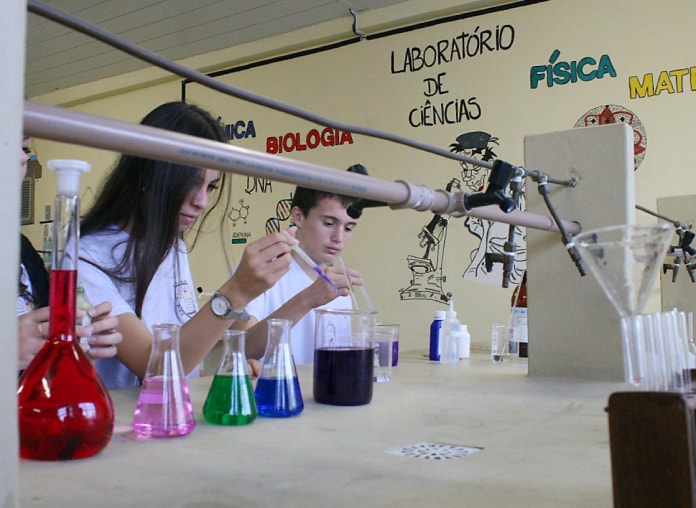 aluno e aluna mexem em ampolas e outros frascos de vidro com líquidos de diferentes cores em uma bancada