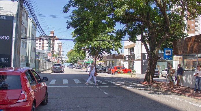 mulher atravessa faixa de pedestre em cruzamento da rua koesa com carro estacionado de um lado e pessoas em pé na calçada do outro