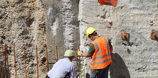dois operários com equipamentos de proteção sobre estrutura de ferro dentro de grande buraco de obra