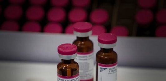 três ampolas de doses de vacina sobre uma bancada e outras organizadas em bandeja ao fundo