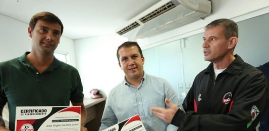 três homens posam para foto, com prefeito no meio com certificado na mão olhando para a câmera