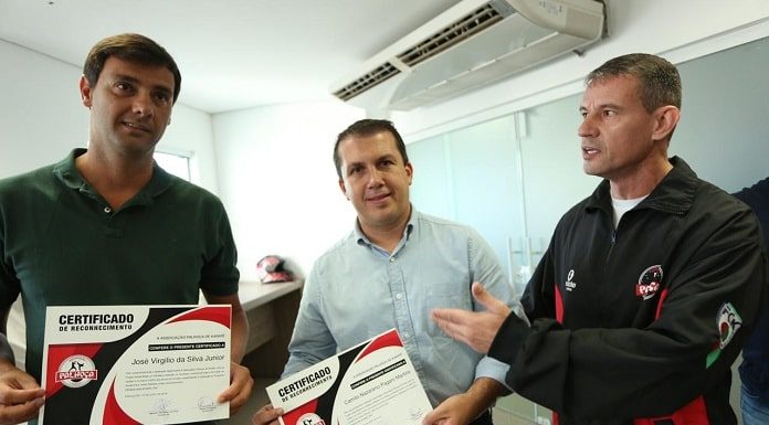 três homens posam para foto, com prefeito no meio com certificado na mão olhando para a câmera